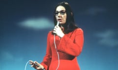 Nana Mouskouri, straight hair past her shoulders and wearing glasses and a wide-sleeved dress, holds a microphone to her mouth as she sings and holds the cord with her other hand
