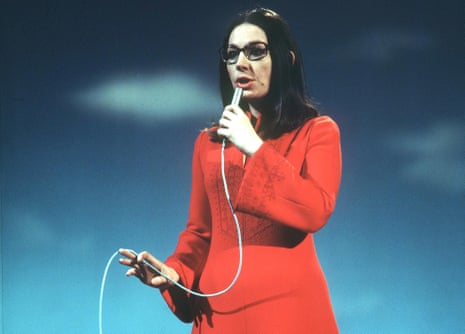Nana Mouskouri, straight hair past her shoulders and wearing glasses and a wide-sleeved dress, holds a microphone to her mouth as she sings and holds the cord with her other hand