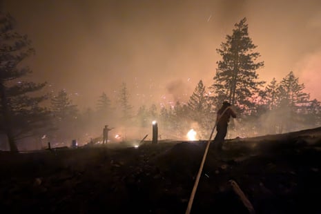 Firefighter silhouetted against burning trees, dragging a hose