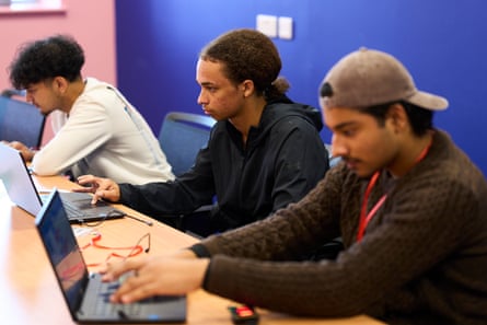 Three teenage boys sit at a long table looking intently at laptop screens. The wall behind them is painted deep blue.