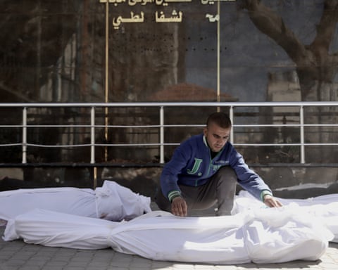 A man crouches between shrouded bodies