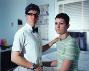 A same-sex couple in a bedroom, one with their hand on the other's hip and the other with their hand on the other's shoulder, both facing the camera with clear, serious expression
