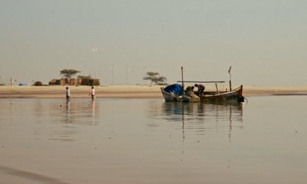 A still from a film shows a boat on calm sea next to a beach. Two people are on the boat, another two wade through the water