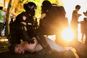 Two police in riot gear hold a bear-chested man face down on the ground