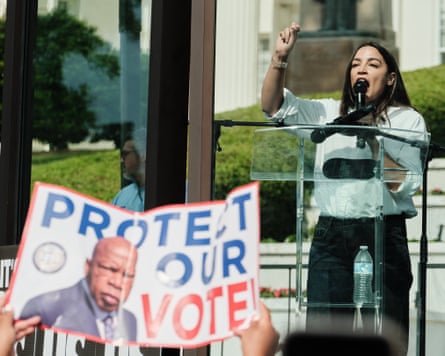 Woman in white top and black pants speaking at lectern while gesturing, as someone in foreground holds poster that reads "Protect Our Vote".