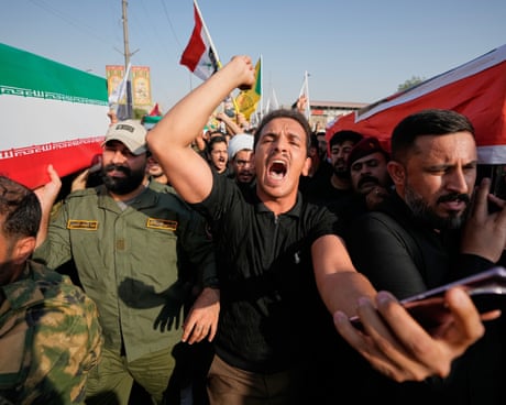 A mourner with open mouth and raised arm walks between men bearing coffins draped with Iranian and Iraqi flags