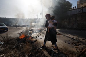 Jalazone, West Bank: A woman carrying a child walks past a burning tyre after Israeli forces killed Palestinians in refugee camp, in the Israeli-occupied West Bank. The Israeli military says it was undertaking a raid early Monday morning and alleges the two suspects tried to ram their car into soldiers, a claim that could not be independently verified