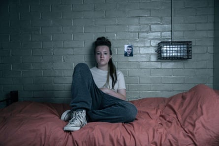Bella Ramsey sitting with her back to a painted brick wall on prison bed, wearing blue jeans, a white T-shirt and white high-top trainers