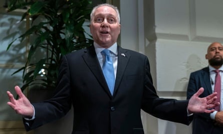 A round balding middle-aged white man with white hair and a dark suit, speaking with both arms and hands outstretched.