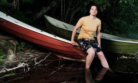 Micheala Bailey, in a T-shirt and shorts with a floral pattern, sits on the edge of a boat with her feet in the water of a lake