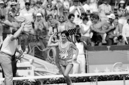 A black and white image of a female athlete waving an American flag as a crowd watches in the background.