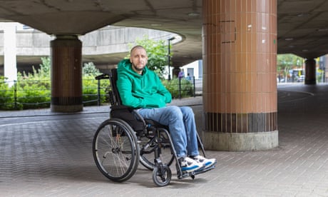 Adam Gabsi in a hoodie, jeans and trainers sitting in a wheelchair next to pillars in an underpass