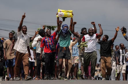A crowd of people pointing, waving and raising a fist march along a road, one of them holding up a small banner in support of Issa Tchiroma