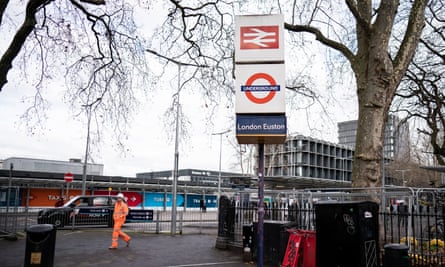Мъж в костюм с висока видимост, вървящ по бордюра на натоварен път до знак, показващ стария символ на британската железница и символа на метрото