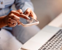 Close-up of hands typing on a smartphone beside an open laptop keyboard