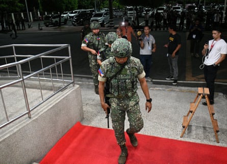 Uniformed troops in camouflage and helmets, carrying guns, walk into the building as people outside look on.