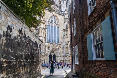 A narrow lane with stone wall on one side and an old stone building on the other with blue wooden shutters by its windows opens out to give a view of York Minster.
