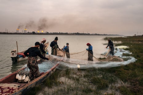 Six men by two small boats prepare their nets where the water meets the marshy shallows. Gas flares from chimneys on the horizon