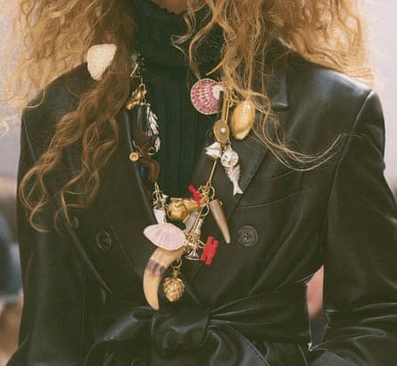 A model with curly hair and round sunglasses wears a leather coat draped with charm necklaces
