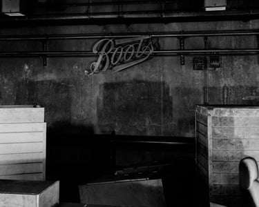 A lit cursive “Boots” sign hangs on a grimy wall above wooden crates in a dim warehouse