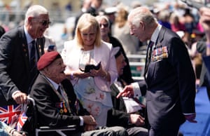 King Charles III in blue jacket with lots of medals meets a second world war veteran wearing red beret and medals in a wheelchair