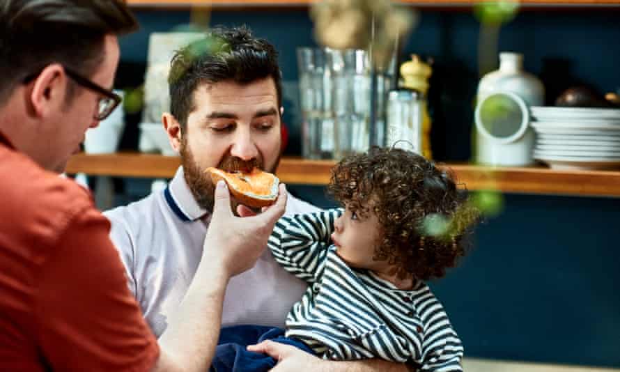 Male couple sharing a bagel with son at home.