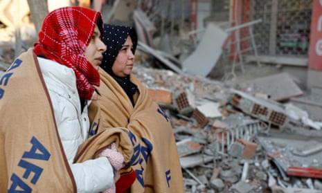 Women wrapped in blankets look on amid rubble in the aftermath of a deadly earthquake in Kahramanmaraş.
