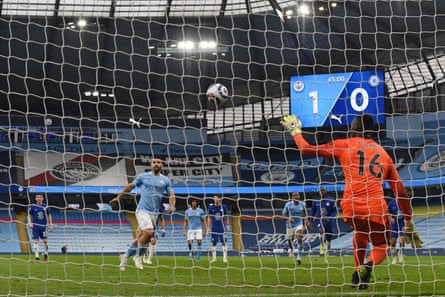 The Chelsea goalkeeper Édouard Mendy stops his dive to the right in time to stop, change direction, and save the Manchester City striker Sergio Agüero’s Panenka penalty. The stadium, whose big screen shows Manchester City 1-0 Chelsea, is almost empty due to the Covid-19 pandemic.