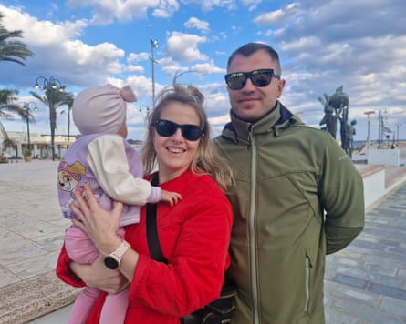 Karin and Oliver Kiilaspa, Karin holding their baby daughter, smile as they stand outside wearing sunglasses in Ayia Napa