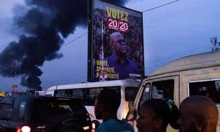 Pedestrians and minibuses pass by an election billboard with the slogan in French saying: ‘Unity, security, prosperity’. A plume of black smoke rises in the background