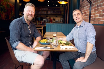 Miles and Celestino both facing the camera and smiling while sat at their table.