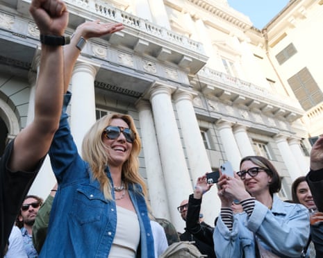 In sunglasses, a white vest and blue denim shirt, Silvia Salis, the mayor of Genoa, waves to a surrounding crowd