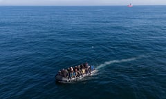 Aerial view of about two dozen people sitting on the edges of an inflatable dinghy motoring across the sea with a large ship in the background