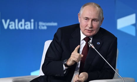 Vladimir Putin extends a finger as he leans towards a microphone on stage in front of a backdrop advertising Valdai Discussion Club.