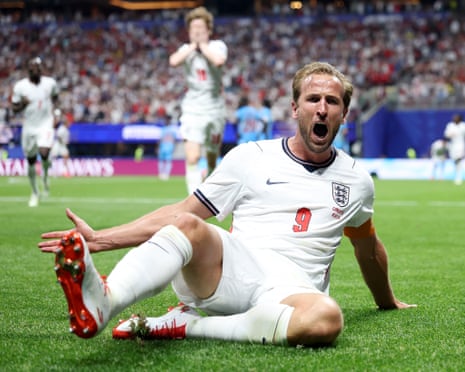 Harry Kane in white England kit sits on the pitch celebrating with his mouth open and arm outstretched