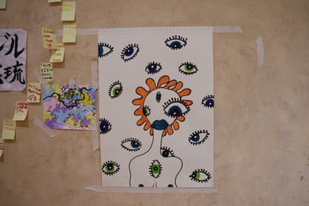 A picture of a figure with orange, petal-like hair, green lips and a number of differently-sized eyes surrounded by green eyes, is taped to a piece of hardboard wall. BNExt to it are notes on post-its.