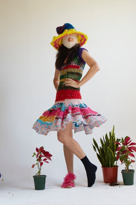 A model in a multi-coloured sleeveless crochet dress with a above-knee flare skirt. She is also wearing a crochet sunhat, a crochet balaclava, a pink sock, and a black sock. Various pot plants are arranged on the floor.