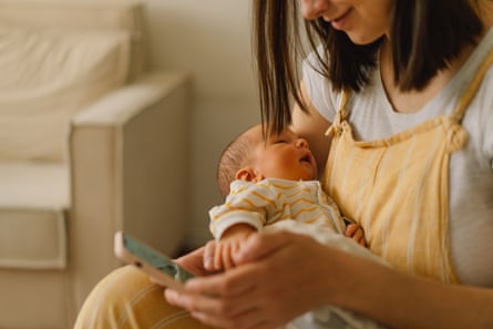 A woman sitting on a bed and typing on a phone while her baby is napping