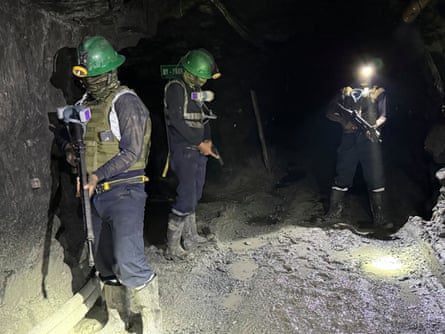 Three masked men in bullet-proof vests and miners’ helmets hold semi-automatic guns in a tunnel