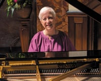 Susan Tomes, a woman with short grey hair and wearing a purple shirt, sits at a grand piano and smiles at the camera