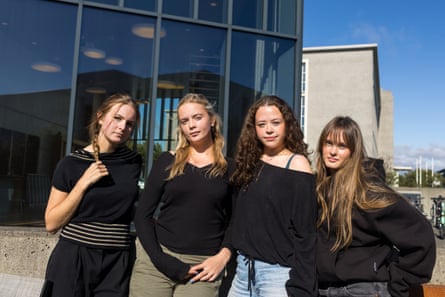Four young women outside a modern glass-fronted building, all with long hair and wearing black tops over jeans or trousers. Saga has very blond hair with a fringe and wears a baggy hoodie, while Anna María, who is also blond with her hair tied back from her face, wears a V-necked jumper over khaki trousers.