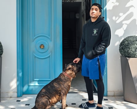 A man in a cap, hoodie, shorts, tight sports troursers and sliders pets a dog standing outside external double doors painted duck egg clue