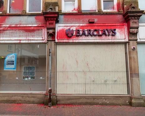 A closed Barclays bank branch storefront splattered with red paint on its signage, shutters, and pavement