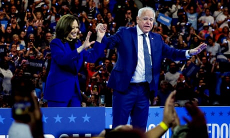 a woman (Kamala Harris) and a man (Tim Walz) in blue suits stand on stage and wave at the crowd