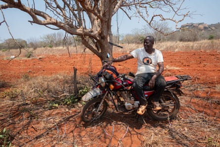 A man in a white T-shirt and black trousers sitting on a motorbike under a tree