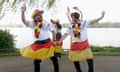 Two Indian men dressed in hats, leis and flags in the German colours of black, red and gold, dance by the side of a lake with two similarly dressed women dancing behind them