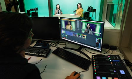 A woman sits in front of a computer screen, with two more women visible in a small TV studio against a green background