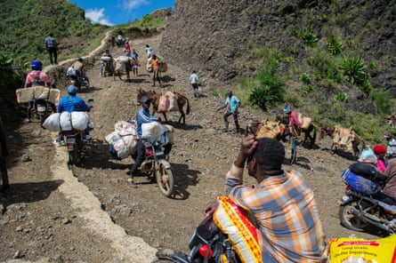 Haitians cary sacks of rice and bundles in tarpaulins up a steep track on motorbikes or donkeys