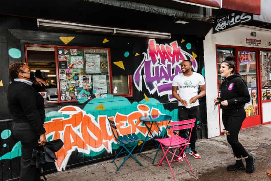 A woman places her order at a walk-up window at a small business with graffiti identifying it as Maya Halal. Nearby a man and woman stand smiling.