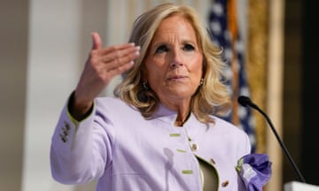 Older white woman with blond hair and lavender suit with purple scarf in the pocked gestures with one hand as she stands at a microphone.